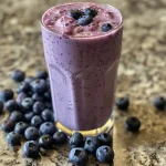 Close-up view of a blueberry smoothie in a glass, showcasing rich purple hues and fresh blueberries on top.