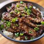 Close-up view of a crispy beef rice bowl garnished with green onions and sesame seeds.