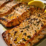Close-up of grilled salmon fillet topped with lemon butter sauce and garnished with lemon wedges.
