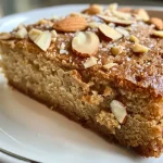 A close-up side view of a moist gluten-free vegan almond cake topped with chopped almonds.
