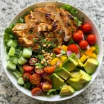Close-up view of a colorful chicken salad bowl filled with greens, tomatoes, and sliced avocado.