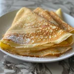 Close-up view of gluten-free crepes stacked on a plate, showing their golden edges.