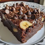 ['Close-up side view of a luscious Double Chocolate Banana Cake topped with chocolate chips.', 'A slice of Double Chocolate Banana Cake, showcasing its rich chocolate layers and banana pieces.', 'Juicy and decadent Double Chocolate Banana Cake with visible chocolate chips and a moist texture.', 'Side view of a homemade Double Chocolate Banana Cake highlighting its chocolatey exterior and garnish.']