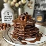 ['Close-up of gluten-free chocolate pancakes drizzled with a rich sauce and topped with hazelnuts.', 'Side view of decadent gluten-free chocolate pancakes, showcasing their fluffy texture and chocolate sauce.', 'Juicy chocolate pancakes stacked high, glistening with a creamy Ferrero Rocher-style sauce.', 'Delicious gluten-free chocolate pancakes with a drizzle of sauce and sprinkled hazelnuts, served on a plate.']