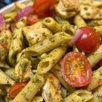 ['Close-up shot of a vibrant Chicken Pesto Pasta Salad featuring penne, chicken, and colorful tomatoes.', 'Side view of a delicious Chicken Pesto Pasta Salad, showcasing fresh ingredients and a creamy pesto dressing.', 'Juicy Chicken Pesto Pasta Salad with halved tomatoes and chopped red onion, captured in a close-up format.', 'Detailed view of a bowl filled with Chicken Pesto Pasta Salad, highlighting the texture of the pasta and ingredients.']