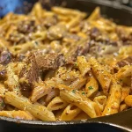 ['A plate of creamy BBQ ranch steak pasta topped with melted cheese and fresh herbs.', 'Close-up view of a savory one-skillet steak pasta dish, featuring rich creamy sauce.', 'Juicy strips of steak mixed with pasta in a creamy barbecue ranch sauce in a skillet.', 'A colorful serving of penne pasta coated in a thick creamy sauce with pieces of steak.']