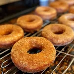 ['Close-up view of baked apple cider donuts with a golden brown crust and a sugary topping.', 'Side view of freshly baked apple cider donuts, showcasing their fluffy texture and cinnamon sugar coating.', 'Juicy apple cider donuts with a glossy finish and sprinkled with a mix of granulated sugar and spices.', 'Delicious baked apple cider donuts, displaying rich colors and a tempting outer layer of sugar.']
