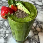 ['A close-up side view of a creamy green smoothie in a glass with fresh ingredients around.', 'A refreshing green smoothie with spinach and avocado, displayed next to chia seeds and strawberries.', 'A vibrant green smoothie in a clear glass, featuring chunks of avocado and surrounded by fresh berries.', 'Smoothie in a glass showing a creamy texture with spinach and colorful strawberries nearby.']