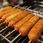 ['Close-up view of crispy mini corn dogs on a wooden platter.', 'Side view of golden mini corn dogs skewered on wooden sticks.', 'Juicy mini corn dogs arranged neatly with a dipping sauce.', 'A cluster of mini corn dogs showcasing their crispy exterior.']