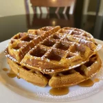 ['Close-up view of golden pumpkin waffles topped with syrup and spices.', 'Side view of a stack of pumpkin waffles drizzled with maple syrup.', 'Juicy pumpkin waffles displayed with a sprinkle of cinnamon and maple syrup.', 'Deliciously fluffy pumpkin waffles served on a plate with syrup glistening.']